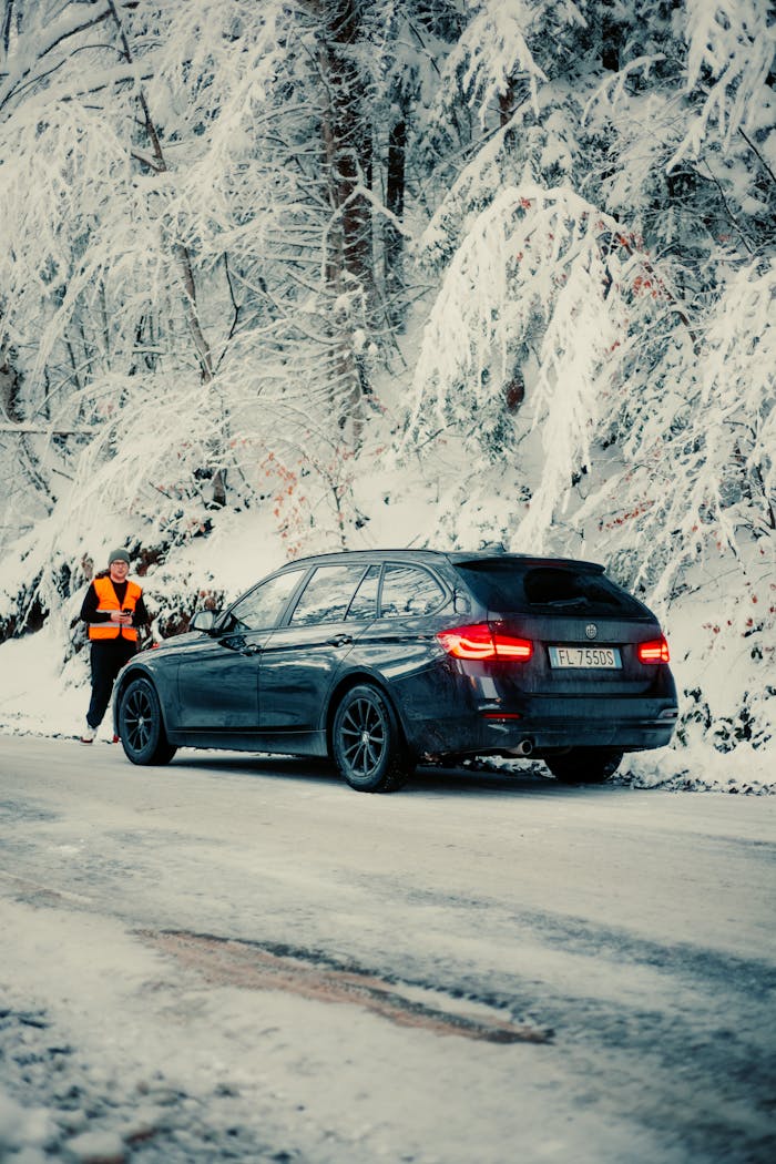 A person assists a vehicle stuck on a snowy road in a winter forest.